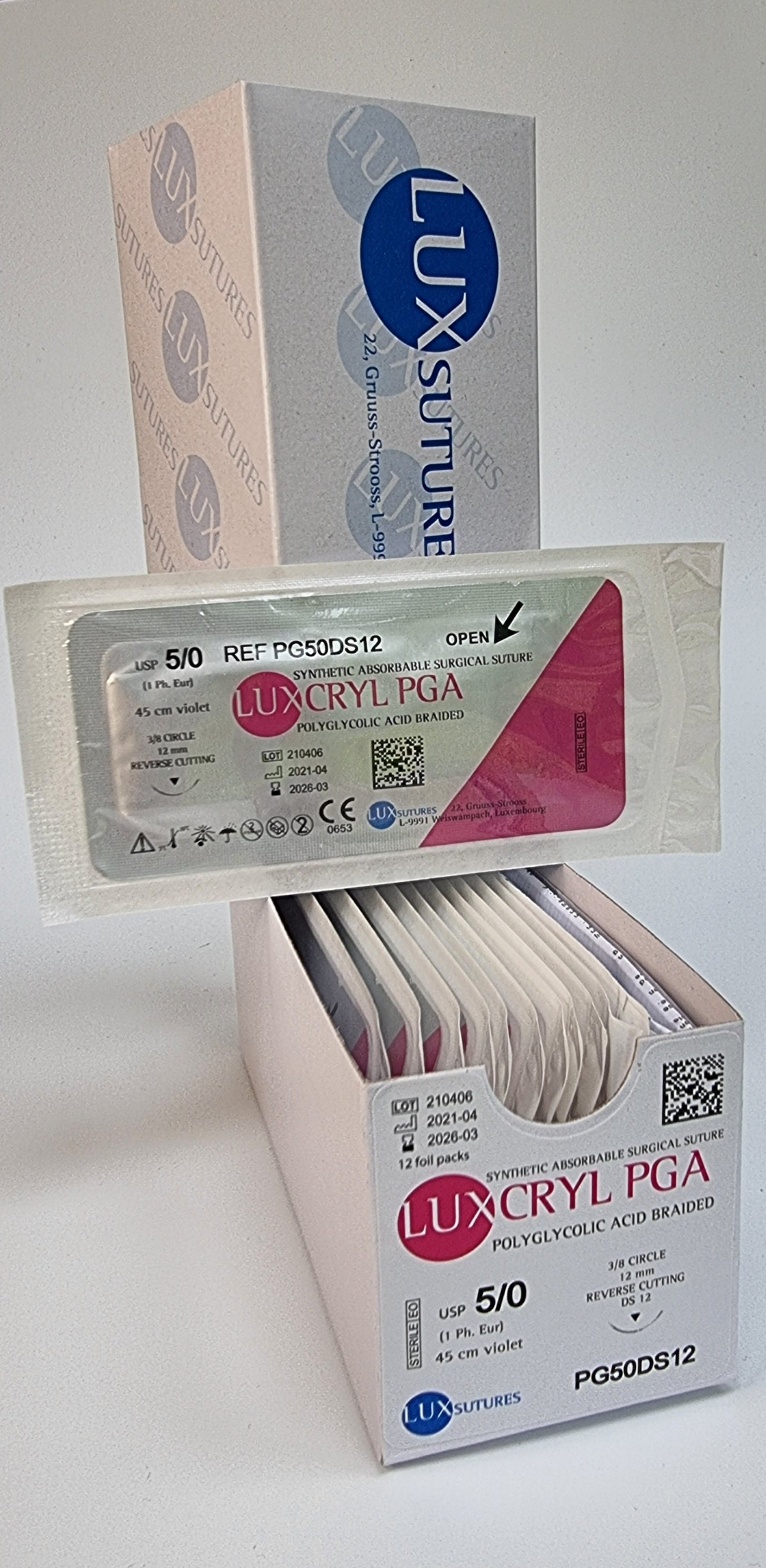 A box of Luxcryl PGA synthetic absorbable surgical sutures is displayed, with one individually packaged suture shown in front. The label indicates USP 5/0 (1 Ph. Eur.), 45cm violet suture, and a 12mm 3/8 circle reverse cutting needle. The packaging highlights the product as polyglycolic acid braided sutures, with the lot number, manufacturing, and expiration dates clearly visible. The sterile packaging is emphasized against a plain background.