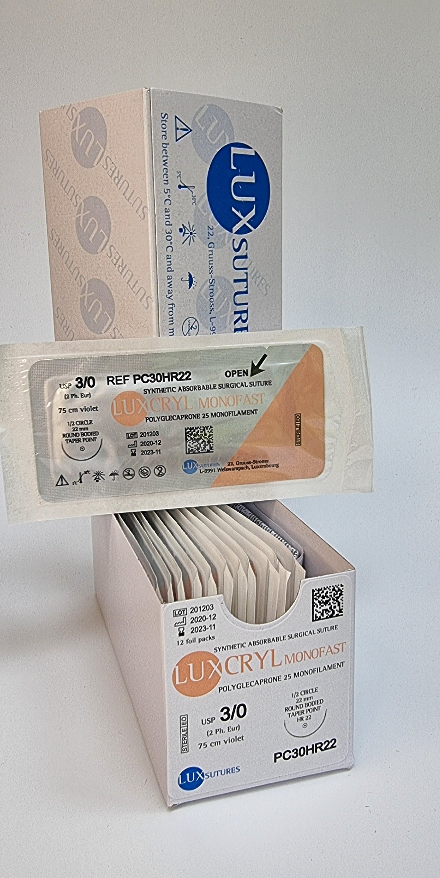 A box of Luxcryl Monofast synthetic absorbable sutures is displayed, with one individually packaged suture shown in front. The label indicates USP 3/0, 75cm violet suture, and a 22mm 1/2 circle round-bodied taper point needle. The sterile packaging and product branding are highlighted against a plain background.