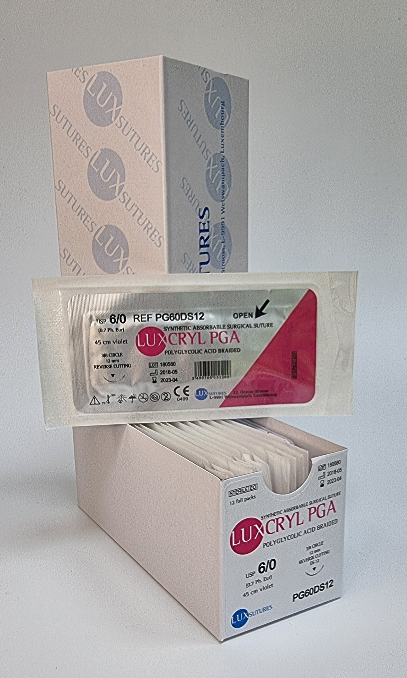 A box of Luxcryl PGA synthetic absorbable sutures is shown, with one individually packaged suture displayed in front. The label specifies USP 6/0, 45cm violet suture, and a 12mm reverse cutting needle. The packaging is white with pink and blue branding, and the product is intended for surgical use. The background is plain, emphasizing the sterile packaging.