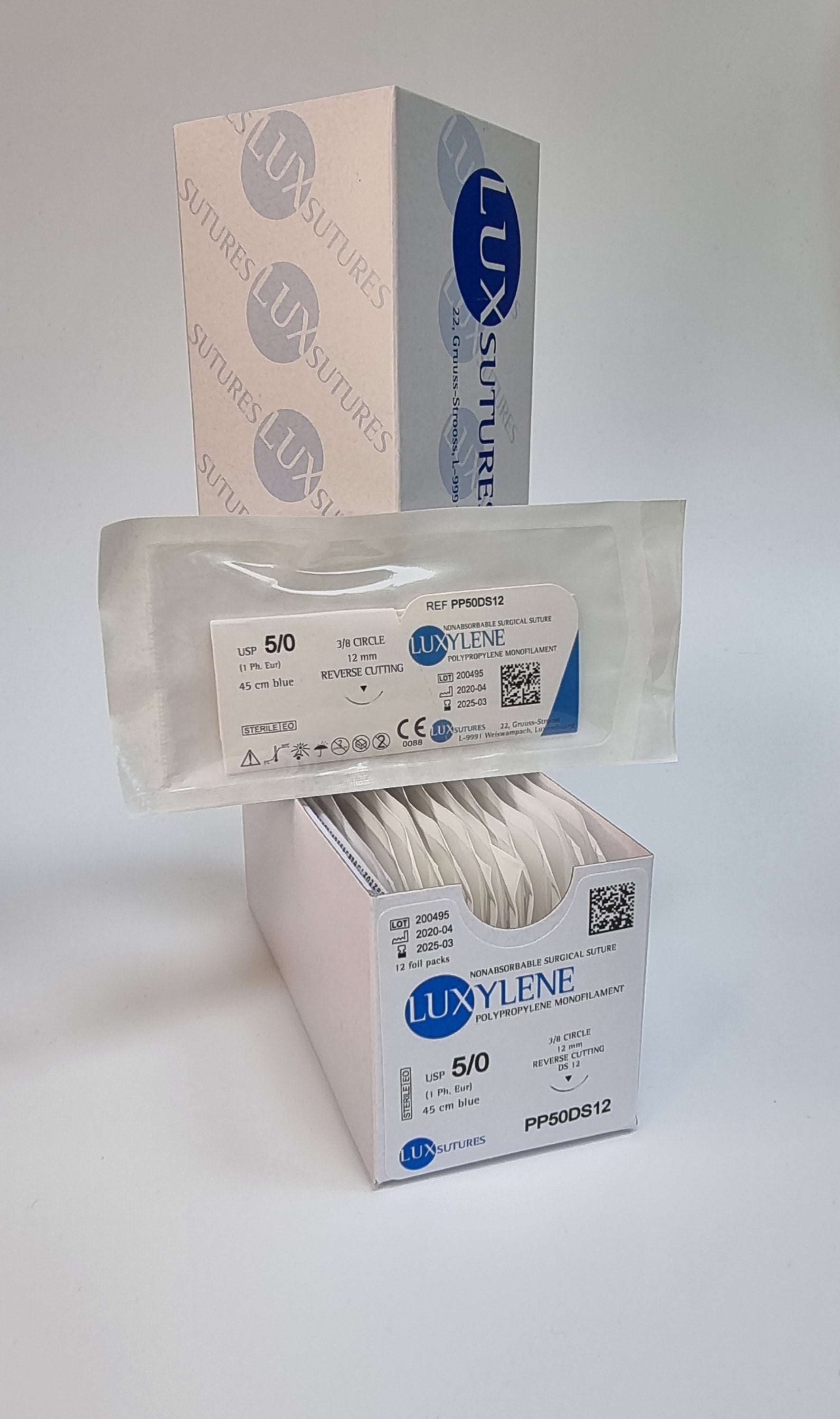 A box of Luxylene polypropylene monofilament non-absorbable surgical sutures is displayed, with one individually packaged suture shown in front. The label indicates USP 5/0, 45cm blue suture, and a 12mm 3/8 circle reverse cutting needle. The sterile packaging highlights product details, with the box branding visible in the background.